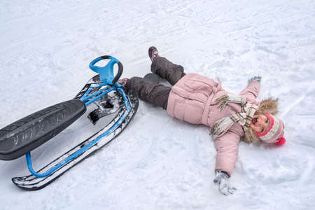 A little girl with a smile and an opened mouth is lying on her back in snow, together with her...