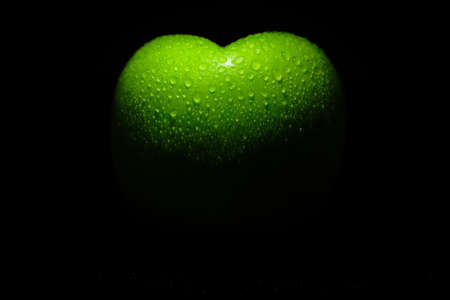Green apple in the shape of a heart with water drops on a black background.の写真素材