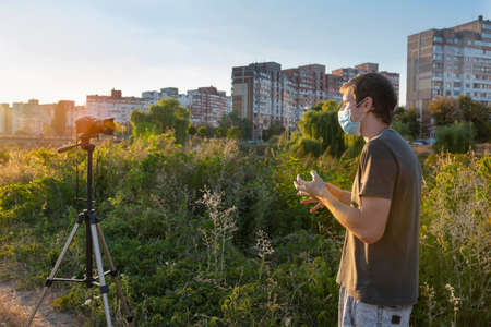 A young man in a medical mask records a video on the camera at sunset. Online communication, consultations, training, businessの写真素材