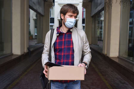 A sad business man in a protective mask with a box of things in his hands, against the backdrop of a modern building in the background. Concept of unemployment problem caused by Covid-19 coronavirus.の写真素材