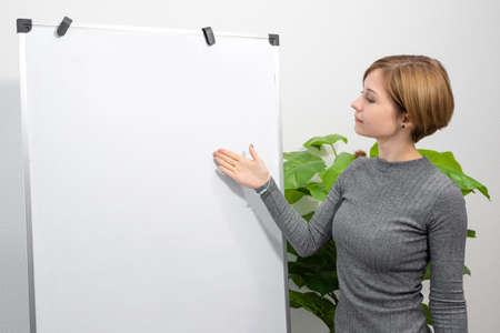 A young woman is standing in a bright room pointing at a blank flip chart. Online teacher, tutor.の写真素材
