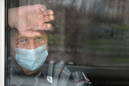 A man in a protective mask in quarantine looks through the window. Quarantine, self-isolation, coronavirus pandemic outbreak, lifestyle, healthcare concept.の写真素材