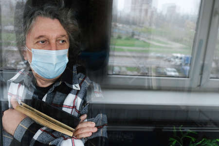 An elderly man in a protective mask in quarantine at home, looking out the window. Self-isolation...