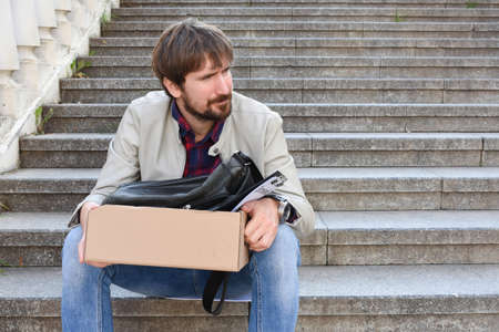 Unemployed man sitting on the stairs outdoors with a box of things. Fired a frustrated desperate man. Unemployment conceptの写真素材
