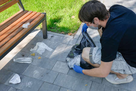 A male volunteer with rubber gloves removes garbage and household waste in a trash bag. Public park cleaning during quarantine. Environmental problems. Environmental protection concept.の写真素材