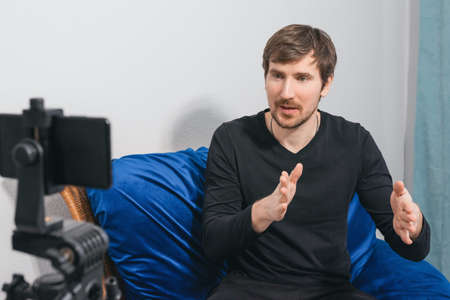 Portrait of a talking male psychologist sitting on the couch and recording a video blog at home. A...