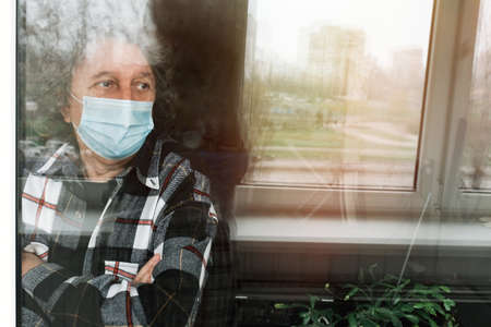 An elderly man in a protective mask on self-isolation at home, looks out the window.の写真素材