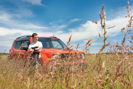 A teenager stands next to a car in a field in dense grass.の写真素材
