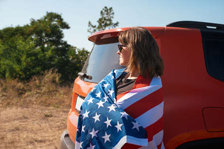 Young woman with American flag in nature near the car. Free lifestyle