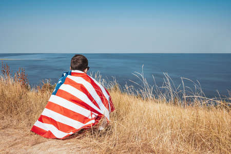 Teenager sits with the American flag on the bay outdoors. Back viewの写真素材