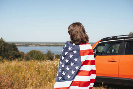 Young woman with American flag standing next to a car in nature. Back viewの写真素材