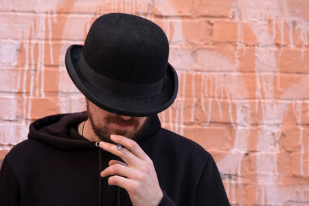 A man in a black hat smoking a cigarette outdoors against a brick wallの写真素材