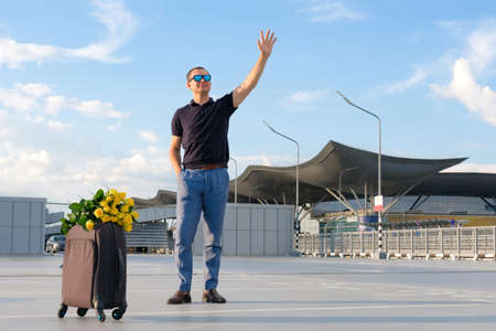 Young man in an airport parking lot with luggage waving to passengers. Business trip, transport, travel and peopleの写真素材