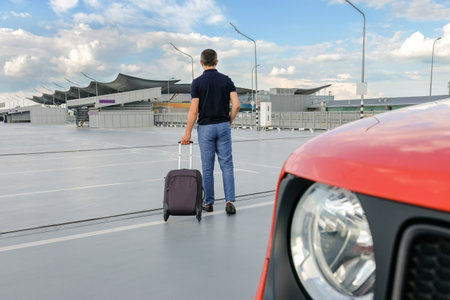 A young man with a suitcase in his hand in the open parking lot of the airport. View from the backの写真素材