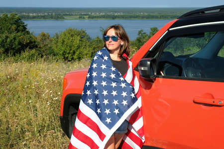 Young woman with america flag stands near car and enjoys nature. Free lifestyle concept.
