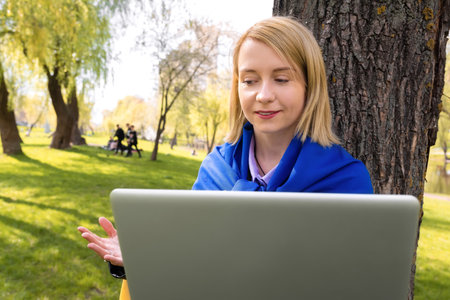 Smiling young Ukrainian woman talking to camera on laptop in park. Video conferenceの写真素材