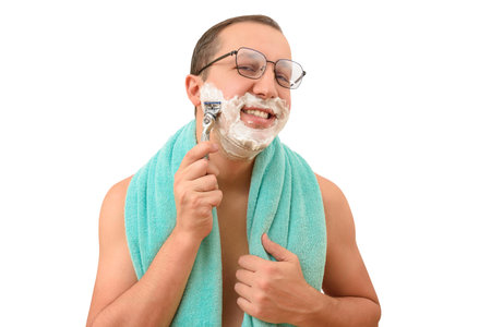 A man with foam on his face and a towel on his shoulders shaves with a razor looking at the camera on a white background. Body care and male hygiene conceptの写真素材