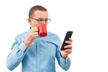 Young man in a shirt drinking a hot drink and looking at a mobile phone on a white backgroundの写真素材
