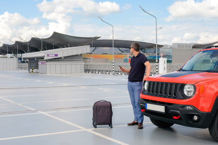 A man with luggage stands near his car in an open airport parking lot.