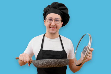 Happy cook with a frying pan in his hand looking at the camera on a blue background. Cooking and people conceptの写真素材