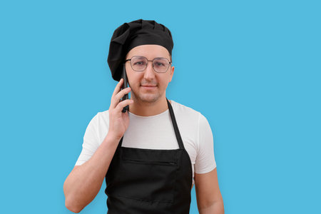 Young chef talking on the phone and looking at the camera on a blue background. The concept of cookingの写真素材