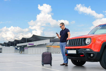 A young man uses a smartphone while standing near a car in an open parking lot. Road lifestyle conceptの写真素材