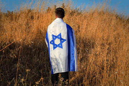 Jewish Israeli young man stands in a field with a large flag of Israel on his shoulders and back. Patriotic holiday concept for Independence day Israelの写真素材