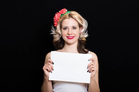 Woman with blonde curls and red lipstick at the rim with red flowers holding a white sheet on a black background and smiling. Close-up. Space for textの写真素材