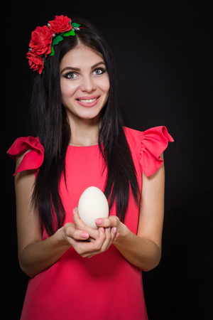 A beautiful girl in a pink dress and a wreath of red flowers is holding a wooden egg.の写真素材