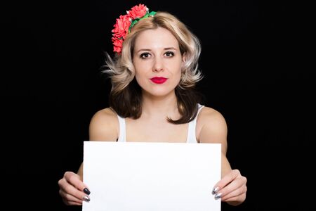 Woman with blonde curls and red lipstick at the rim with red flowers holding a white sheet on a black background and smiling. Close-up. Space for textの写真素材