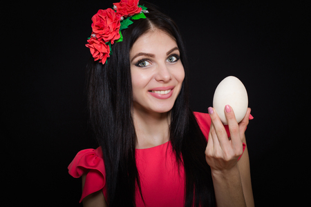A beautiful girl in a pink dress and a wreath of red flowers is holding a wooden egg.の写真素材