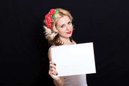 Woman with blonde curls and red lipstick at the rim with red flowers holding a white sheet on a black background and smiling. Close-up. Space for textの写真素材