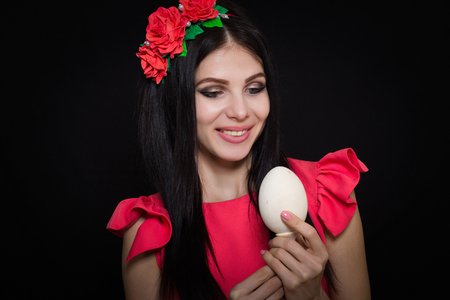 A beautiful girl in a pink dress and a wreath of red flowers is holding a wooden egg.の写真素材