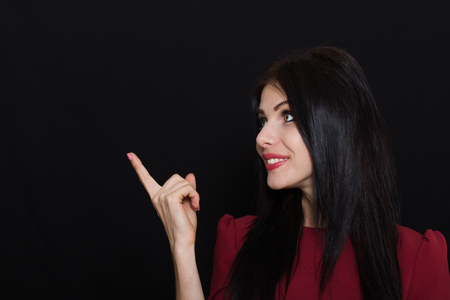 Beautiful woman with black straight hair and blue eyes on a dark background shows her finger and smiles. Close-up. Space for textの写真素材