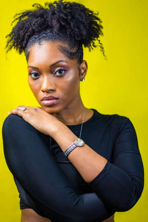 Black woman with natural hair poses with her hand on her shoulder, Black woman wearing silver jewelry poses in front of a yellow backdrop, Portrait of a black woman, Portrait of a woman posingの写真素材