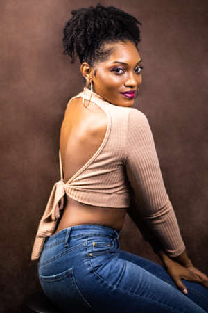 Black woman shows her back wearing brown sweater sitting down posing, Black woman posing in a brown sweater, Woman with natural hair and hoop earrings, Black woman wearing brown sweater and blue jeanの写真素材