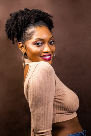 Black woman wearing brown sweater sitting down posing with head turned towards shoulder, Black woman posing in a brown sweater, Smiling woman with natural hairの写真素材