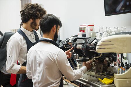 Baku, Azerbaijan, January 12, 2019: Preparing coffee for a client. Handsome young man in apron making coffee at the coffee machine while standing at cafe.のeditorial素材