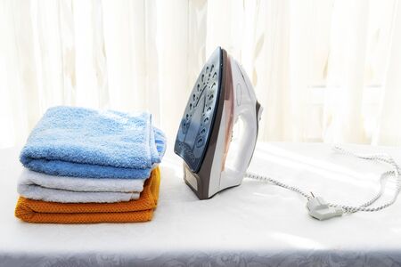 White iron and multicolored towels on the Ironing Board on the background of the windowの写真素材