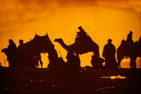 A group of camels and their riders arriving at the top of a sand dune are silhouetted against a dramatic cloudy desert sunset. A distant mountain peak rises above the horizon in the background view.の写真素材