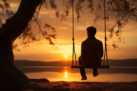 Silhouette of a man sitting on a swing by the lake at sunsetの素材