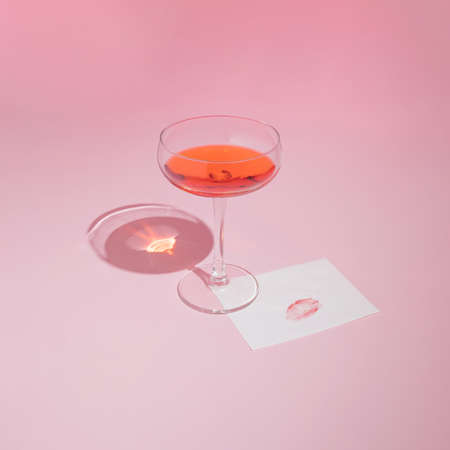 Red wine glass and romantic card with lipstick kiss on pastel pink background. Trendy Valentine's Day aesthetic.の写真素材