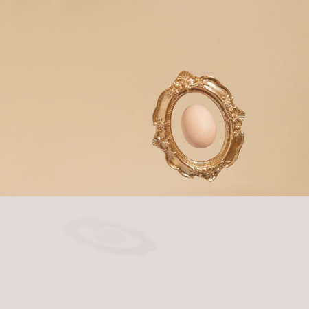 Easter egg framed in vintage baroque style golden frame and floating above beige and white background. Modern minimal style with copy space.の写真素材