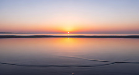 A breathtaking sunset paints the sky in hues of orange, pink, and purple, with the sun's reflection shimmering on the tranquil ocean and wet beach sand.の素材