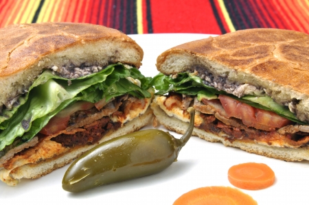 Closeup of halved Mexican torta sandwich with toasted bun and jalapeno pepper on plate over colorful tableclothの写真素材