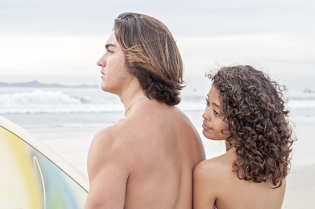 Young attractive man and woman couple stand closely together on gorgeous beach watching surf as he holds surfboardの写真素材
