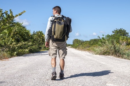 Male hiker with backpack and gear walking alone down lonely asphalt road flanked by tropical...