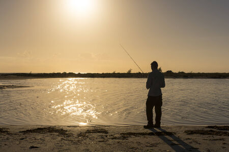 Man fishing in lagoon silhouetted by orange sun over horizonの写真素材