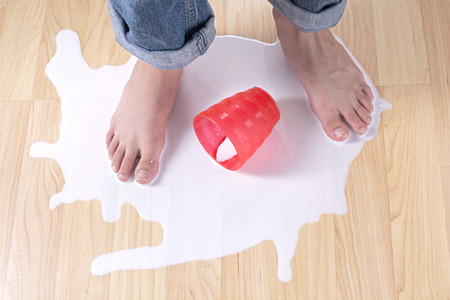 Child feet standing in puddle of spilled milk on wood floor with red cupの写真素材