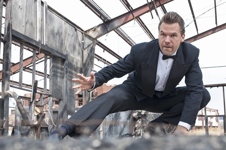 Handsome tough Caucasian man in black tuxedo poses in action stunt scene in destroyed warehouse